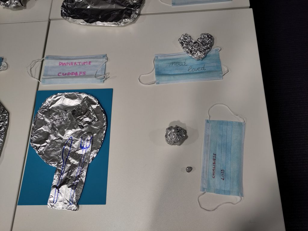 An image of a white table with four pieces of tinfoil art in the shapes of a dinner plate, knife and fork; a love heart, and; a small ball and a slightly larger ball. There are also three blue coloured surgical masks with the words 'dinnertime' and 'cuddles' written on one, 'need' and 'loved' written on the second, and 'loneliness' and 'loss' written on the third.