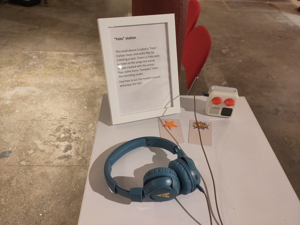 The image shows a photograph of a 'yoto' device - which is used to allow people to listen to audio content. There are blue headphones, a framed image detailing how to use the yoto device, and the small, white yoto device itself. 